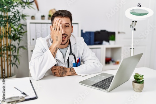 Young doctor working at the clinic using computer laptop covering one eye with hand, confident smile on face and surprise emotion.