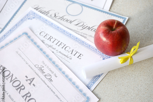 A honor roll recognition, certificate of achievement and high school diploma lies on table with small scroll and red apple. Education documents photo