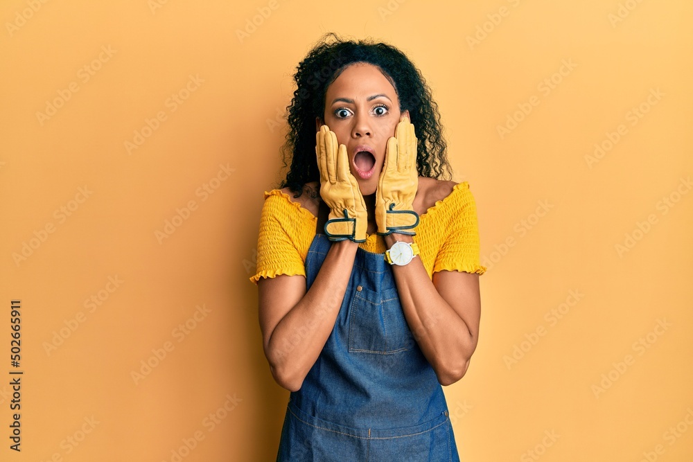 Middle age african american woman wearing professional apron afraid and shocked, surprise and amazed expression with hands on face Middle age african american woman wearing professional apron afraid and shocked, surprise and amazed expression with hands on face