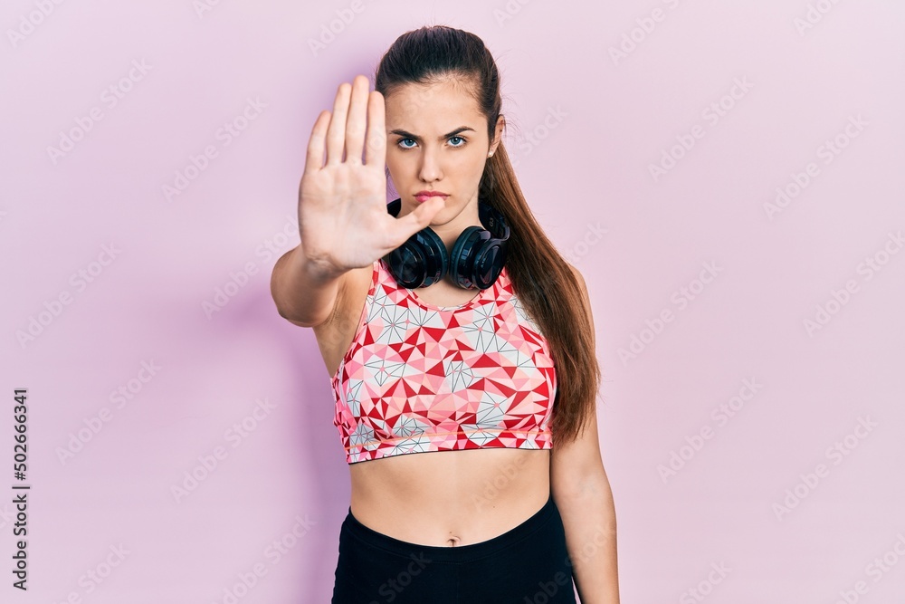 Young brunette teenager wearing gym clothes and using headphones doing stop sing with palm of the hand. warning expression with negative and serious gesture on the face. Young brunette teenager wearing gym clothes and using headphones doing stop sing with palm of the hand. warning expression with negative and serious gesture on the face.