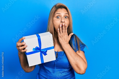 Middle age hispanic woman holding gift covering mouth with hand, shocked and afraid for mistake. surprised expression