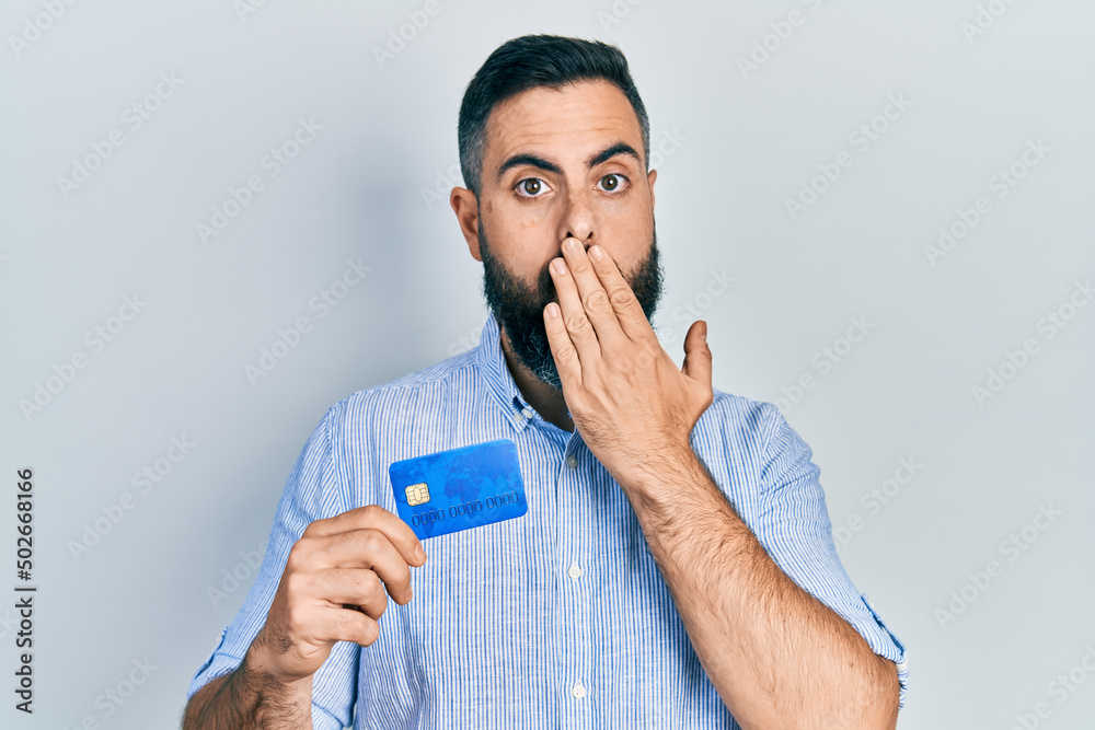 Young hispanic man holding credit card covering mouth with hand, shocked and afraid for mistake. surprised expression