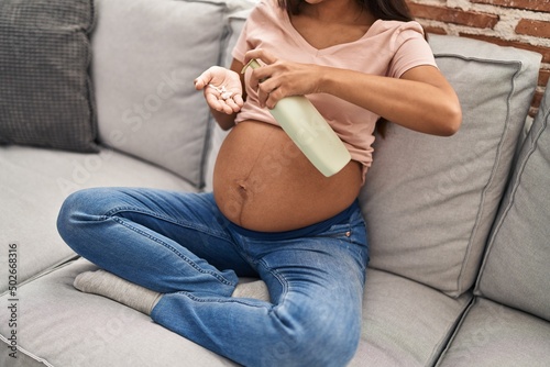 Young latin woman pregnant applying cream lotion on skin belly at home photo