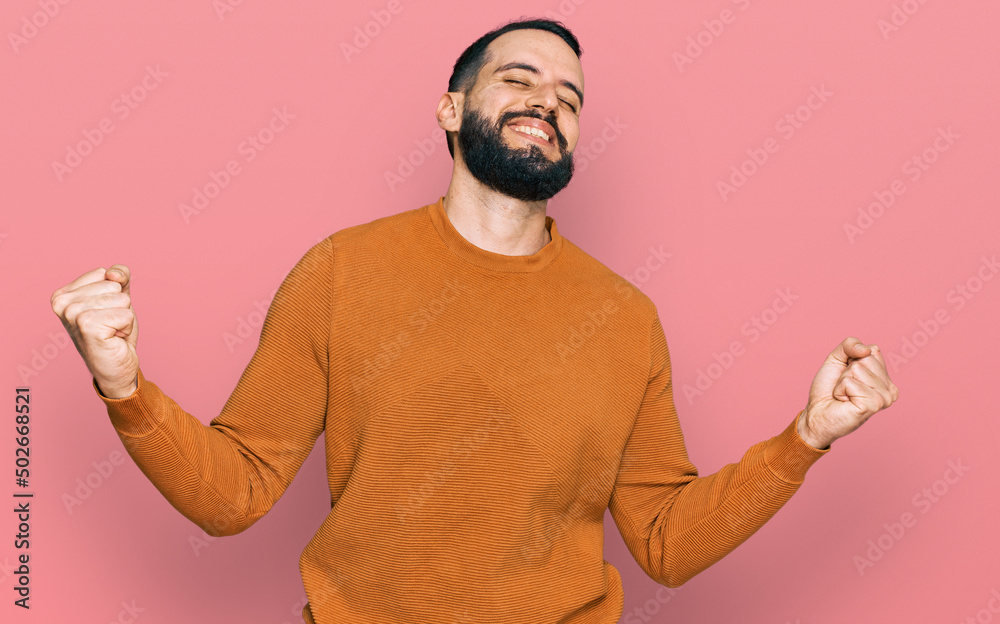 Fototapeta premium Young man with beard wearing casual winter sweater very happy and excited doing winner gesture with arms raised, smiling and screaming for success. celebration concept.