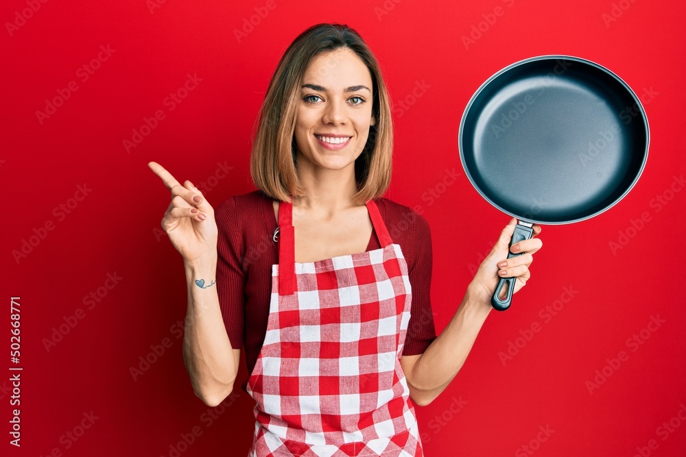 Young caucasian blonde woman wearing cook apron holding pan smiling happy pointing with hand and finger to the side Young caucasian blonde woman wearing cook apron holding pan smiling happy pointing with hand and finger to the side