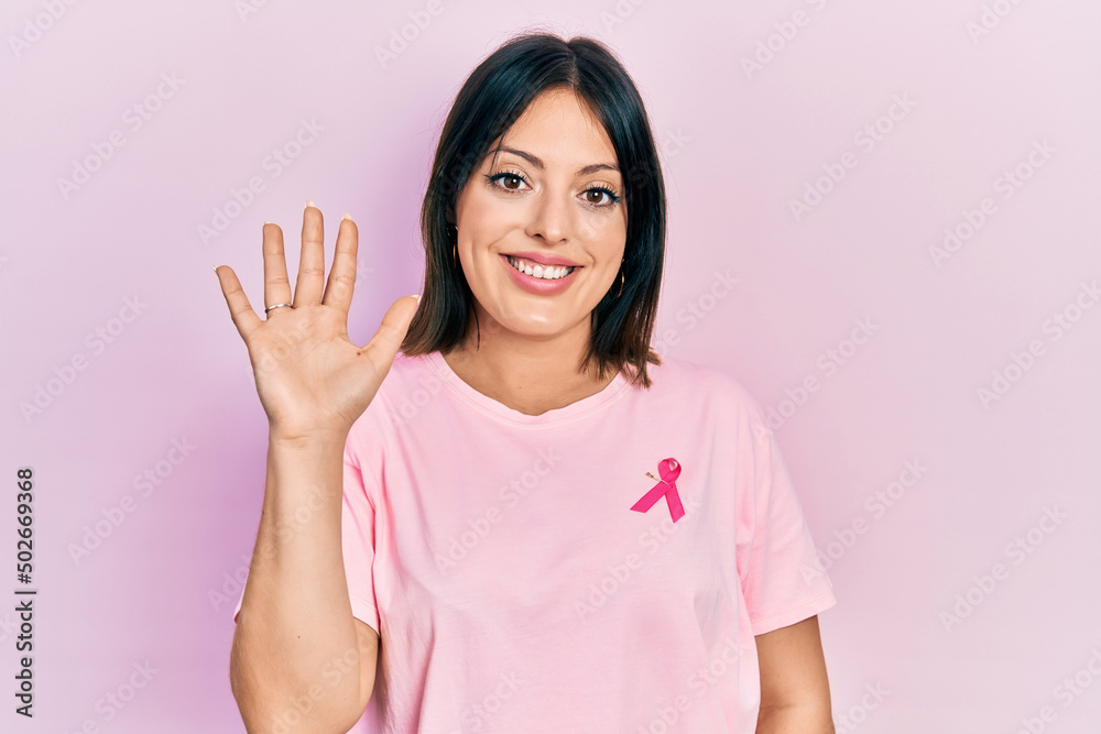 Young hispanic woman wearing pink cancer ribbon on t shirt waiving saying hello happy and smiling, friendly welcome gesture Young hispanic woman wearing pink cancer ribbon on t shirt waiving saying hello happy and smiling, friendly welcome gesture