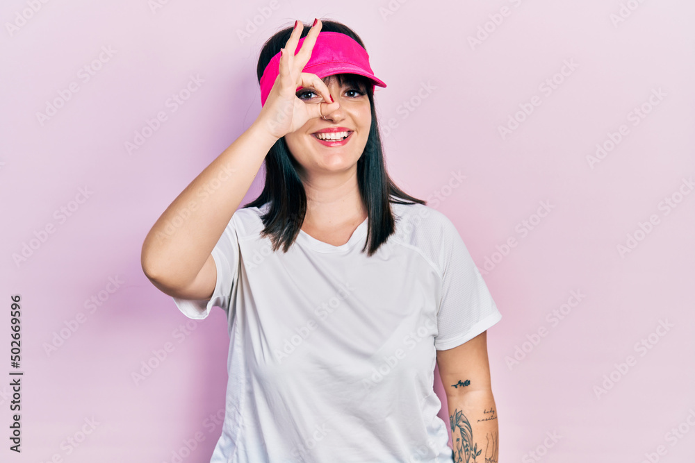Young hispanic woman wearing sportswear and sun visor cap smiling happy doing ok sign with hand on eye looking through fingers Young hispanic woman wearing sportswear and sun visor cap smiling happy doing ok sign with hand on eye looking through fingers