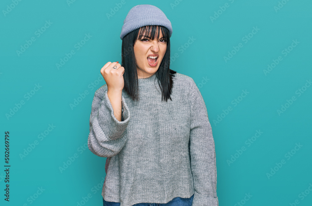 Young hispanic woman wearing cute wool cap angry and mad raising fist frustrated and furious while shouting with anger. rage and aggressive concept. Young hispanic woman wearing cute wool cap angry and mad raising fist frustrated and furious while shouting with anger. rage and aggressive concept.