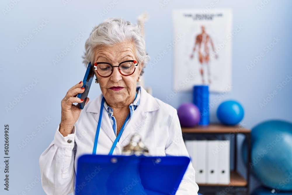 Senior grey-haired woman wearing physiotherapist uniform talking on the smartphone at physitherapy clinic Senior grey-haired woman wearing physiotherapist uniform talking on the smartphone at physitherapy clinic