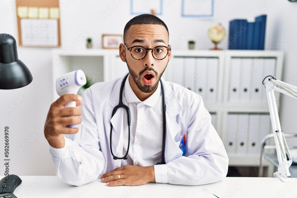 African american doctor man holding thermometer at the clinic scared and amazed with open mouth for surprise, disbelief face African american doctor man holding thermometer at the clinic scared and amazed with open mouth for surprise, disbelief face