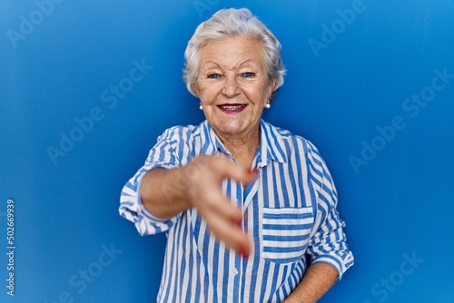 Senior woman with grey hair standing over blue background smiling friendly offering handshake as greeting and welcoming. successful business.