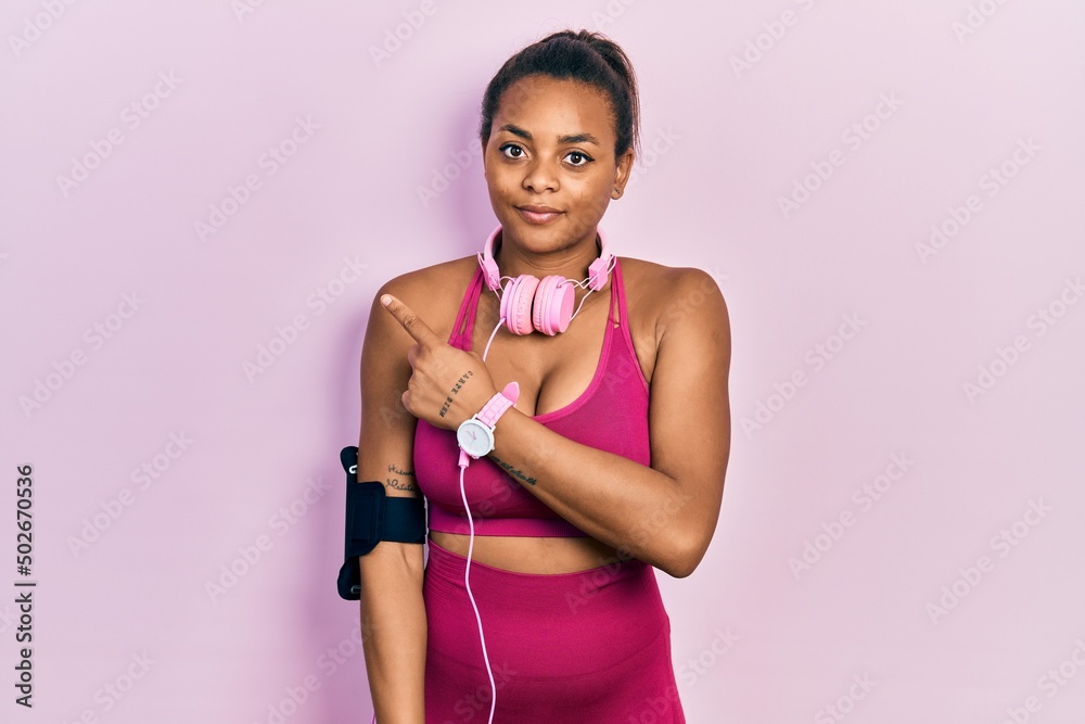 Young african american girl wearing gym clothes and using headphones pointing with hand finger to the side showing advertisement, serious and calm face