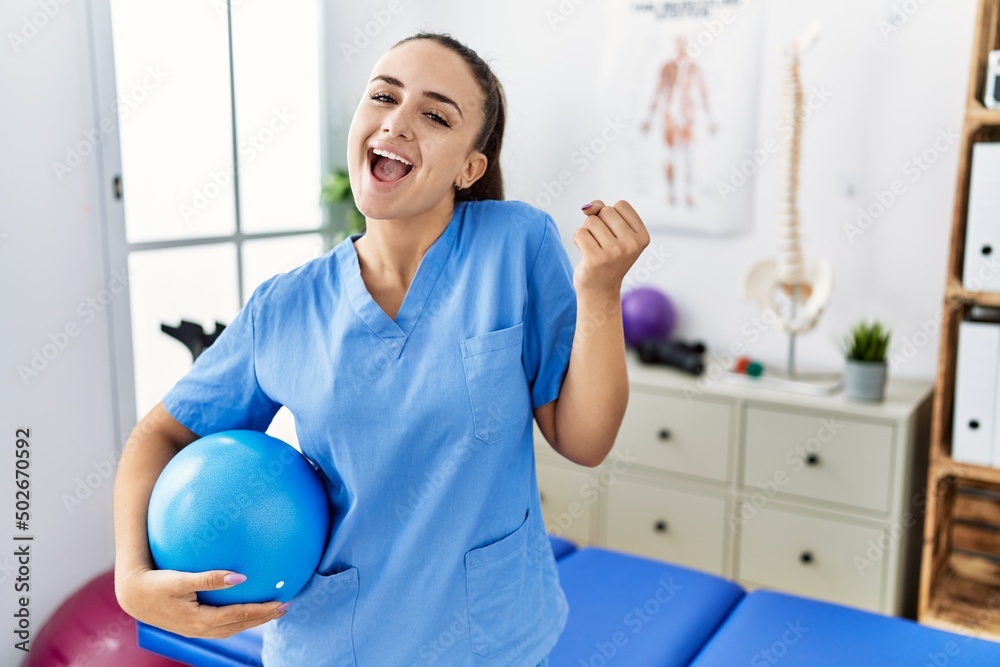 Young physiotherapist woman holding pilates ball at medical clinic screaming proud, celebrating victory and success very excited with raised arms Young physiotherapist woman holding pilates ball at medical clinic screaming proud, celebrating victory and success very excited with raised arms