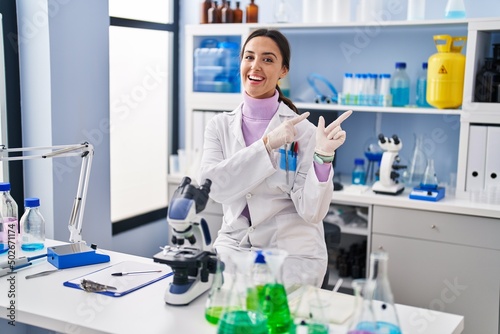 Young brunette woman working at scientist laboratory smiling and looking at the camera pointing with two hands and fingers to the side.
