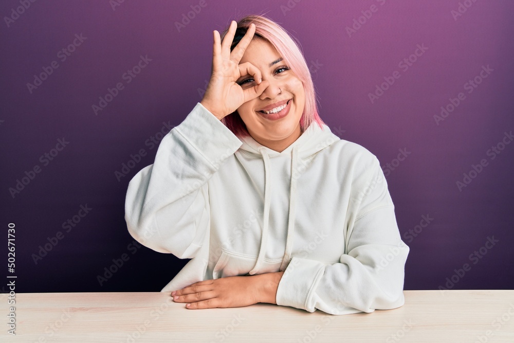 Hispanic woman with pink hair wearing casual sweatshirt sitting on the table doing ok gesture with hand smiling, eye looking through fingers with happy face.