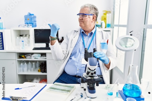 Senior caucasian man working at scientist laboratory smiling with happy face looking and pointing to the side with thumb up.