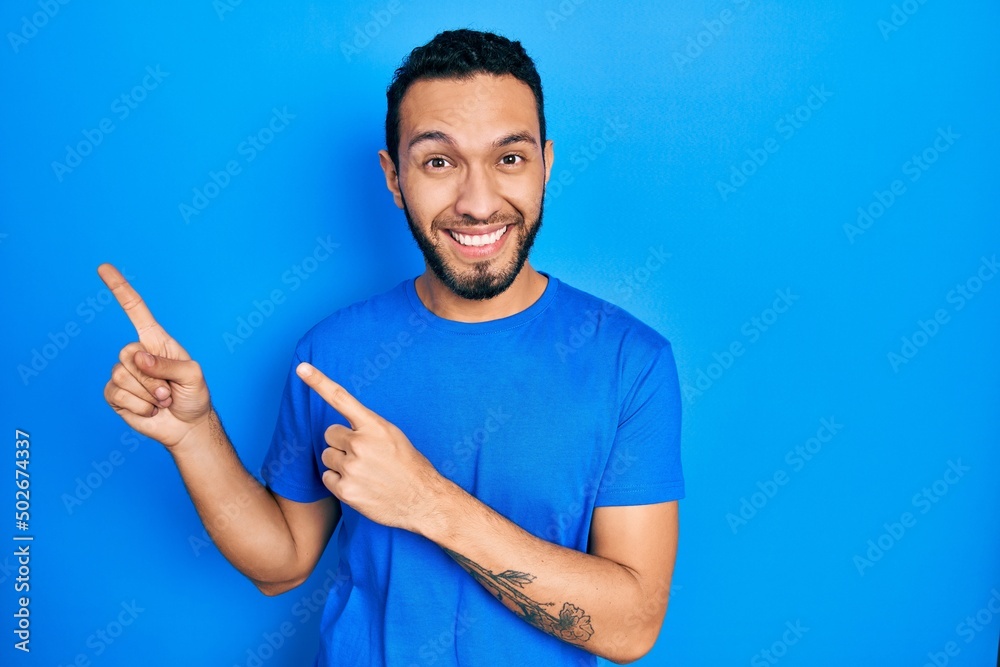 Hispanic man with beard wearing casual blue t shirt smiling and looking at the camera pointing with two hands and fingers to the side. Hispanic man with beard wearing casual blue t shirt smiling and looking at the camera pointing with two hands and fingers to the side.