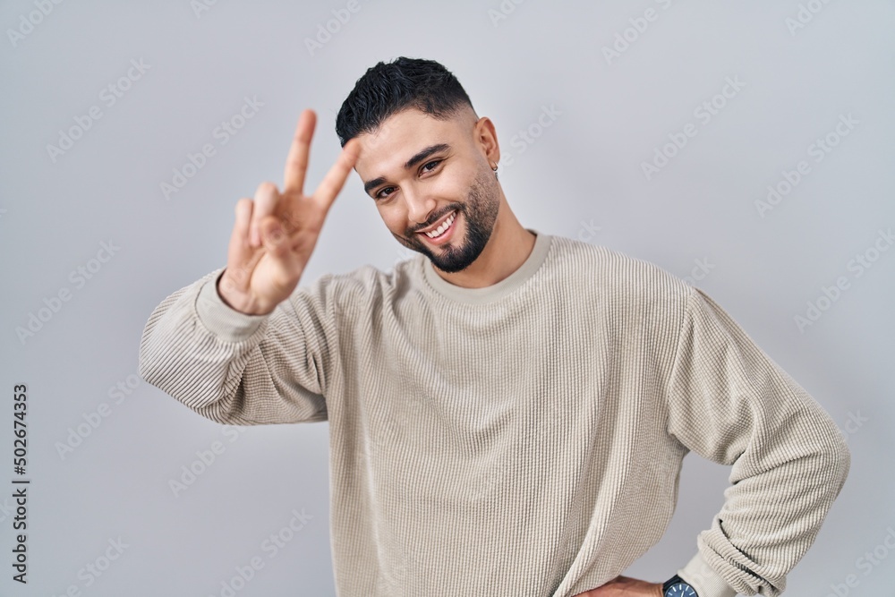 Fototapeta premium Young handsome man standing over isolated background smiling looking to the camera showing fingers doing victory sign. number two.