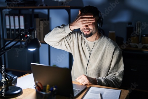 Young handsome man working using computer laptop at night smiling and laughing with hand on face covering eyes for surprise. blind concept.