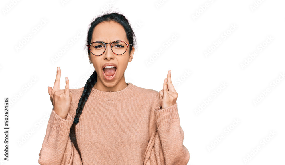 Young hispanic girl wearing casual clothes and glasses shouting with crazy expression doing rock symbol with hands up. music star. heavy concept.