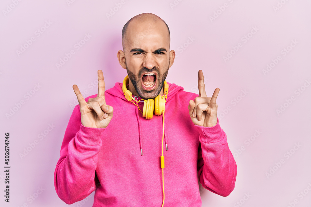 Naklejka premium Young bald man wearing gym clothes and using headphones shouting with crazy expression doing rock symbol with hands up. music star. heavy concept.