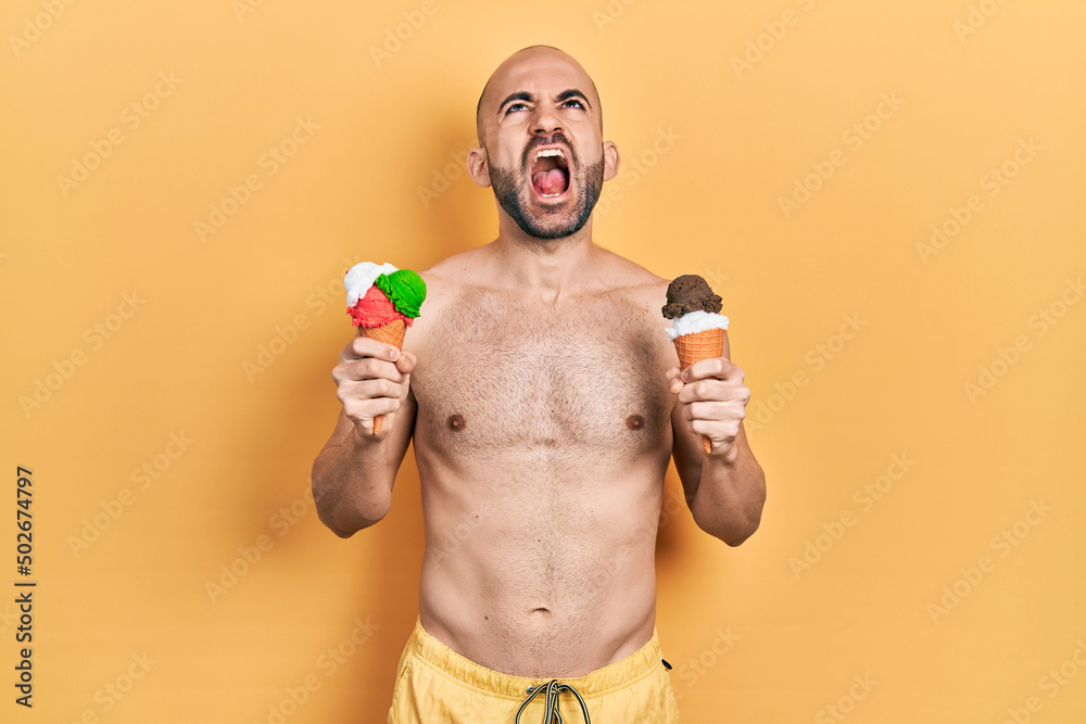 Fototapeta premium Young bald man wearing swimwear holding two ice cream cones angry and mad screaming frustrated and furious, shouting with anger looking up.