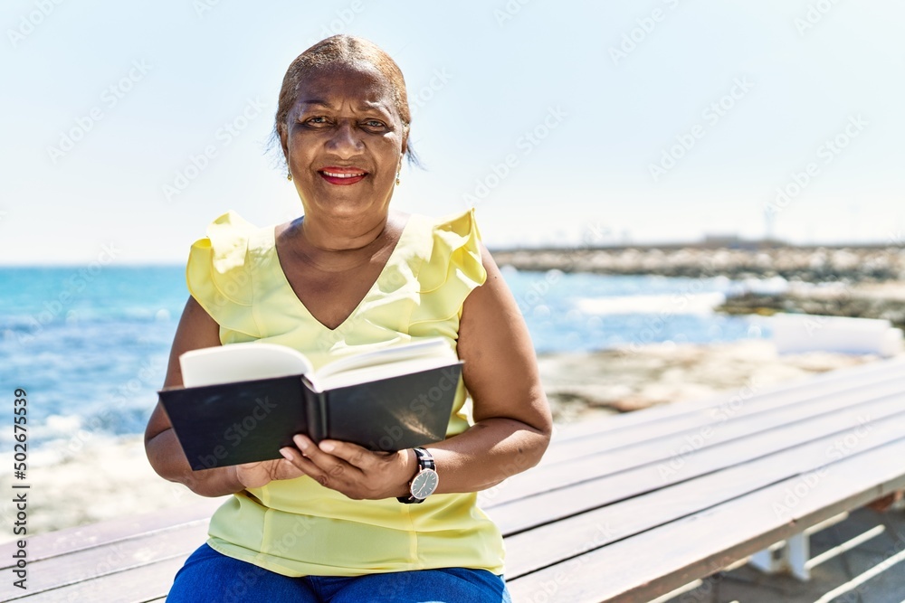 Senior african american woman reading book sitting on the bench at the beach.