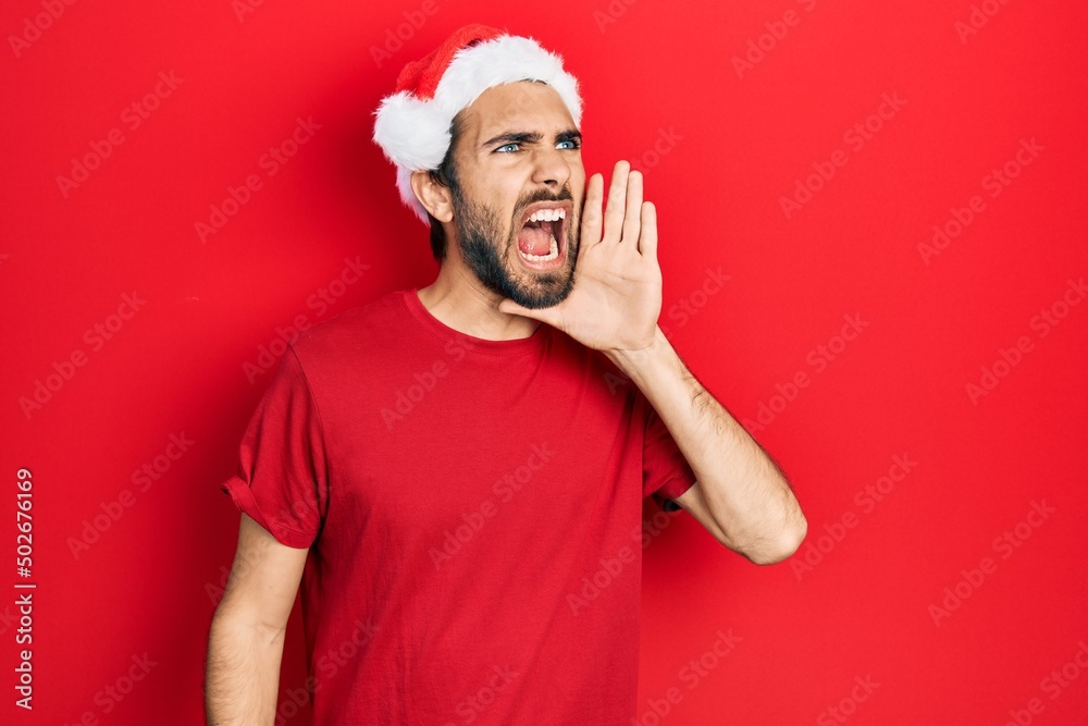 Young hispanic man wearing christmas hat shouting and screaming loud to side with hand on mouth. communication concept. Young hispanic man wearing christmas hat shouting and screaming loud to side with hand on mouth. communication concept.