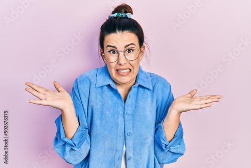 Young hispanic woman wearing casual clothes and glasses clueless and confused expression with arms and hands raised. doubt concept.