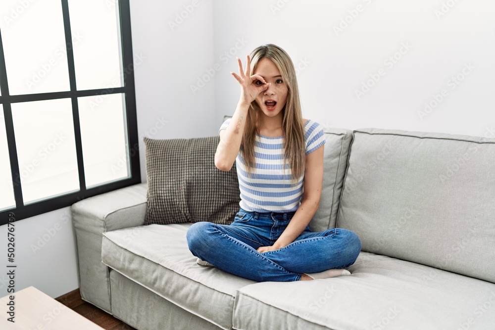Asian young woman sitting on the sofa at home doing ok gesture shocked with surprised face, eye looking through fingers. unbelieving expression. Asian young woman sitting on the sofa at home doing ok gesture shocked with surprised face, eye looking through fingers. unbelieving expression.