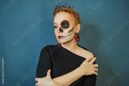 Portrait of a woman with frightening make-up in a black dress with a crown of red beads on a green background. Day of the Dead in Mexico