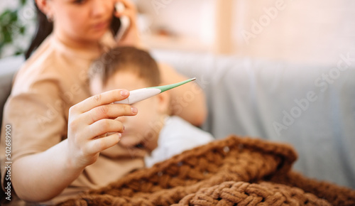 A sad sick boy is lying on the couch, feels fever, flu. The worried mother presses the phone to her ear and calls the family therapist after measuring the child's temperature. selective focus