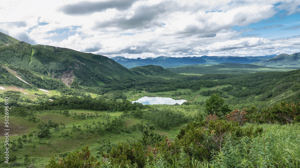 The lake in the valley is surrounded by mountains. There is lush green vegetation on the slopes. The blue sky with clouds is reflected in the water. Kamchatka. Vachkazhets. Lake Tahkoloch