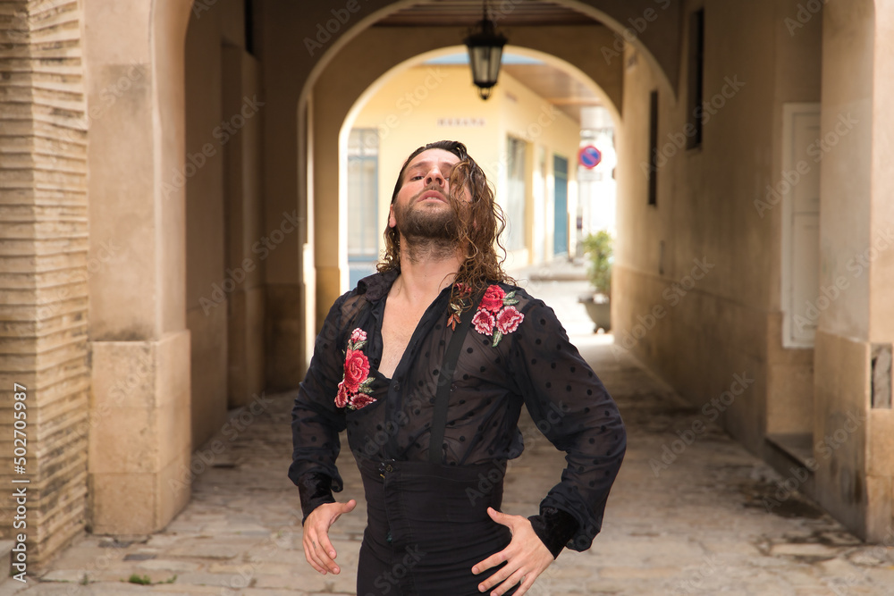 Young man with beard and long hair, wearing black transparent shirt with black polka dots and red roses, black pants and jacket, dancing flamenco in the city. Concept art, dance, culture, tradition.