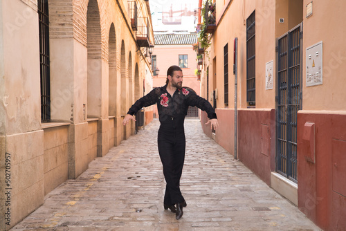 Young man with beard and ponytail, wearing black transparent shirt with black polka dots and red roses, black pants and jacket, dancing flamenco in the city. Concept art, dance, culture, tradition. © Manuel