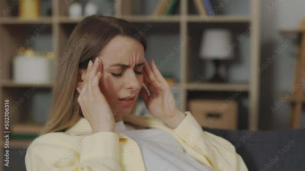Caucasian woman in expectation suffering from migraine while sitting on couch at home. Young future mother massaging temples during nervous tension and stress. Caucasian woman in expectation suffering from migraine while sitting on couch at home. Young future mother massaging temples during nervous tension and stress.