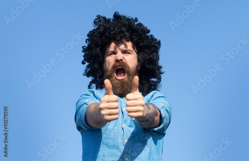 Funny nerdy guy with thumbs up. Crazy funny bearded man with wig on sky background. Expression and success.