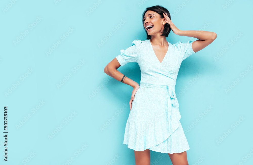 Portrait of young beautiful smiling female in trendy summer dress. Carefree woman posing near blue wall in studio. Positive model having fun indoors. Cheerful and happy. Isolated Portrait of young beautiful smiling female in trendy summer dress. Carefree woman posing near blue wall in studio. Positive model having fun indoors. Cheerful and happy. Isolated