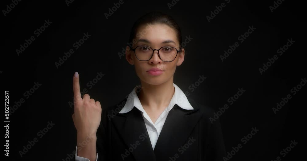 Business woman with glasses in business clothes with a serious face shows one finger with her right hand. Isolated on a black background.
