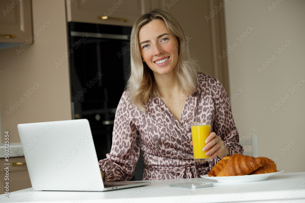 A young smiling girl in a home dress eats croissants and drinks orange juice and works at a laptop