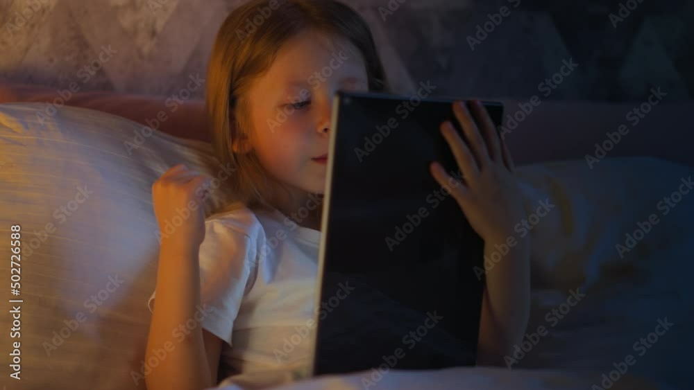 Joyful little girl preschooler happy of winning in videogame expresses joy with raising fist holding modern tablet on bed illuminated by night lamp light in evening slow motion Joyful little girl preschooler happy of winning in videogame expresses joy with raising fist holding modern tablet on bed illuminated by night lamp light in evening slow motion