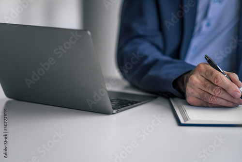 Close-up of senior male executive writing plan in diary by laptop at office desk