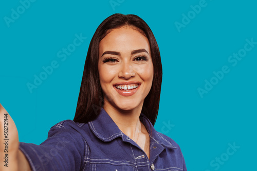 Happy arab woman making selfie. Portrait of smiling girl, posing at blue studio background. Young emotional woman. The human emotions, facial expression concept. Front view.