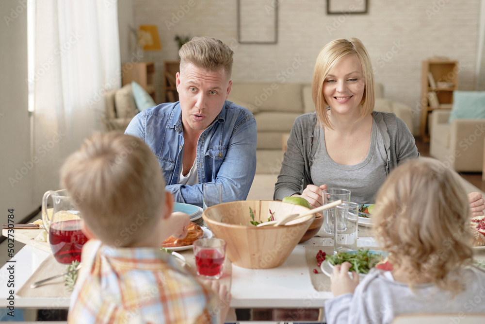 Attractive blond-haired Caucasian couple sitting at dining table with kids, eating appetizing homemade dinner, chatting and enjoying day together