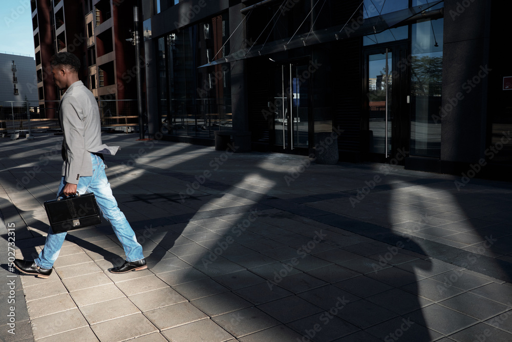 End of working day. Side view of African American manager with case moving away from modern business center, shadows seen on pavement