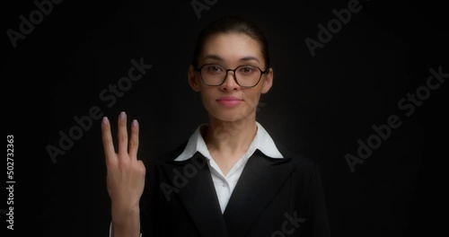 Business woman with glasses in business clothes with a serious face shows three finger with her right hand. Isolated on a black background.