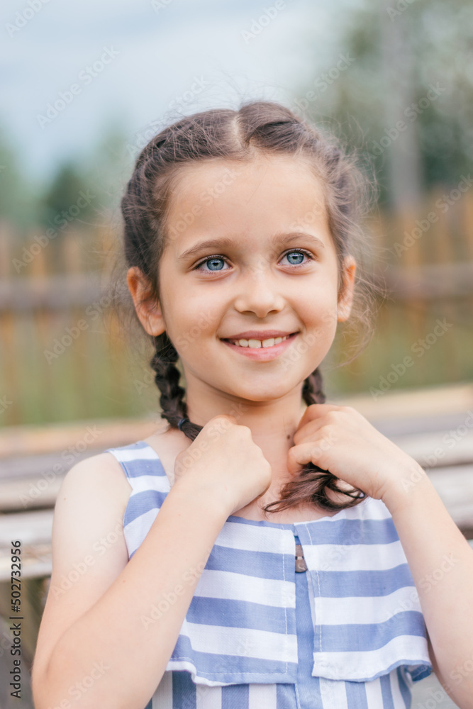 Portrait of a smiling blue-eyed and dark-haired girl with pigtails on the background of a wooden village fence.Childhood, summer, rural simple life, positive vibes. Portrait of a smiling blue-eyed and dark-haired girl with pigtails on the background of a wooden village fence.Childhood, summer, rural simple life, positive vibes.