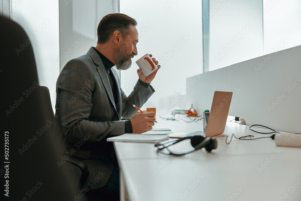 Side view of busy man sitting in modern office while drinking coffee and making notes on documents Side view of busy man sitting in modern office while drinking coffee and making notes on documents