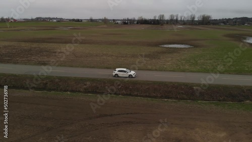 white car 2019 toyota corolla hatchback driving along country farm road surrounded by grass brown fields mountains in farmland Abbotsford BC Aerial wide leading tracking car accelerates side profile
