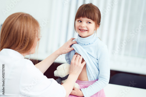 Cheerful female doctor checking patient little girl by stethoscope in hospital